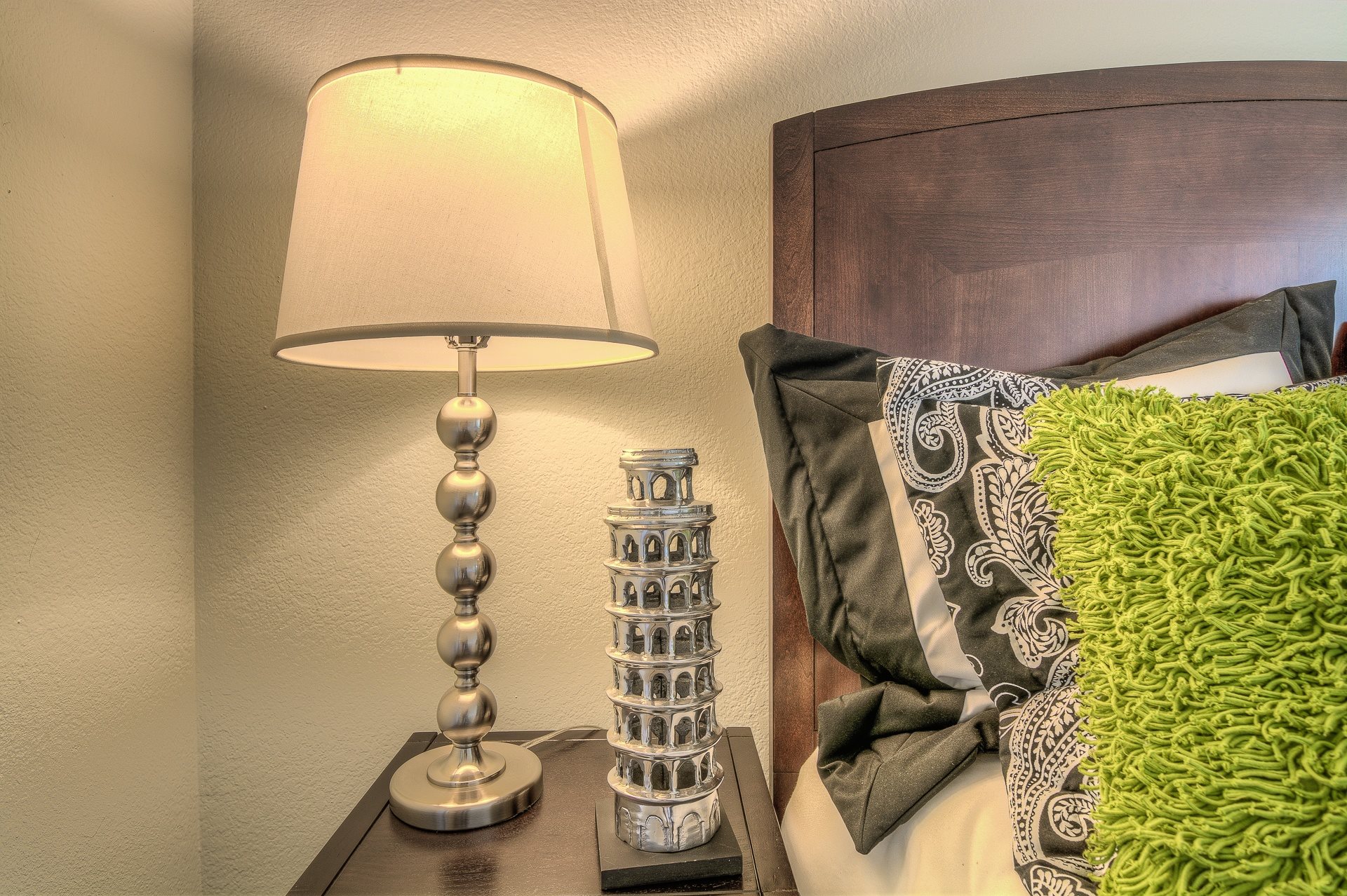 A lamp with a base resembling a tower is on a bedside table next to a bed with a green pillow.,A lamp with a base resembling a tower is on a bedside table next to a bed with a green pillow.